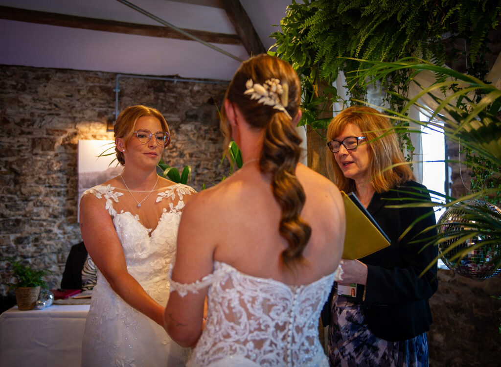 Real Wedding Inspiration - Same-sex wedding ceremony with two brides exchanging vows surrounded by lush greenery and rustic stone walls