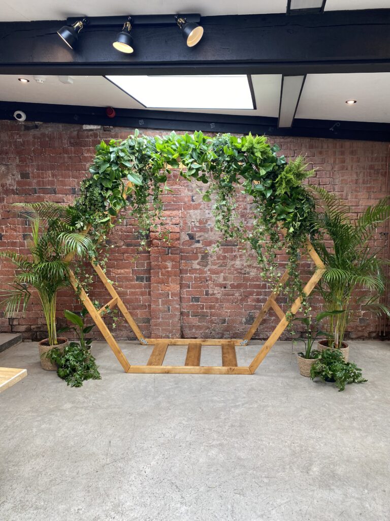 A reclaimed wooden hexagon wedding arch decorated with lush potted greenery and cascading foliage, creating a stunning botanical backdrop for eco-friendly weddings. The structure features living plants such as ferns, palms, and trailing vines arranged to give a natural, garden-inspired feel against a rustic exposed brick wall. Ideal for sustainable or low-waste ceremonies, this living arch combines modern geometric design with vibrant, reusable plant décor — perfect for couples seeking a botanical or eco-conscious wedding style.