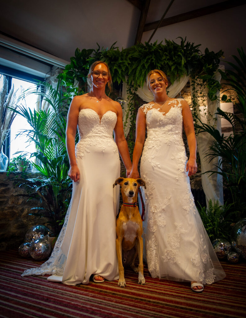 Two brides with their dog standing in front of a lush greenery wedding backdrop