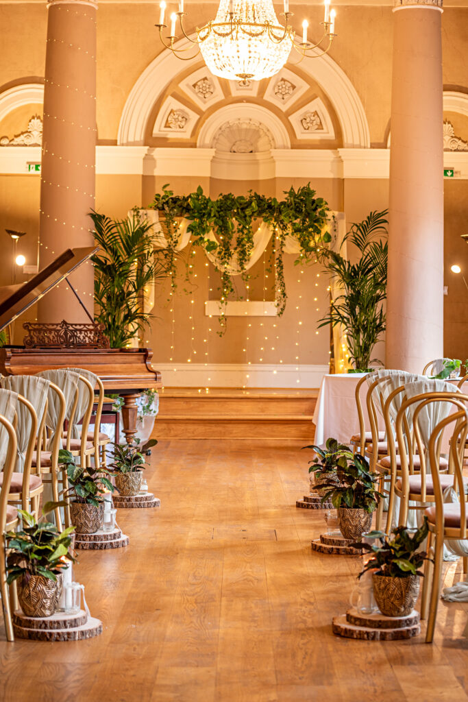 A stunning botanical wedding ceremony setup featuring potted aisle plants, a lush greenery backdrop, and cascading fairy lights. Perfect for eco-conscious couples seeking a sustainable, nature-inspired wedding design with reusable décor and warm, romantic lighting.