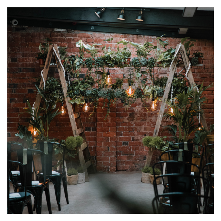 Greenery wedding ceremony backdrop with wooden ladders, hanging plants and warm Edison bulbs against a brick wall