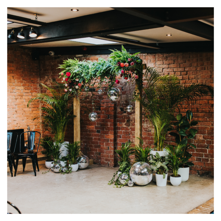 Lush green wedding ceremony backdrop with hanging disco balls, wooden frame and potted plants against exposed brick wall