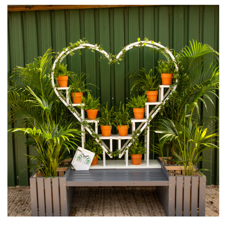 Heart-shaped plant backdrop with potted ferns and palms set against a green outdoor wall.