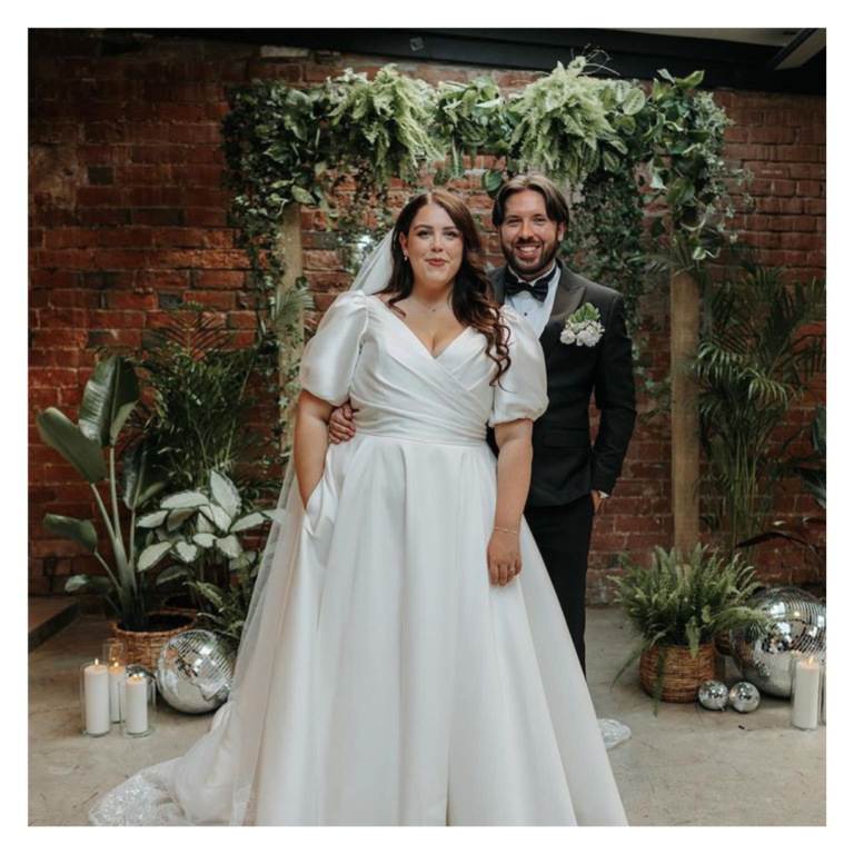 Bride and groom standing together beneath a greenery arch with disco balls and brick backdrop