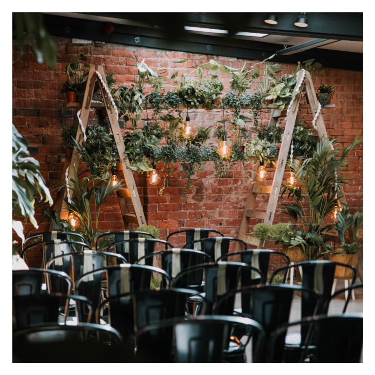 Wedding ceremony seating facing a greenery ladder backdrop with hanging lights and exposed brick wall