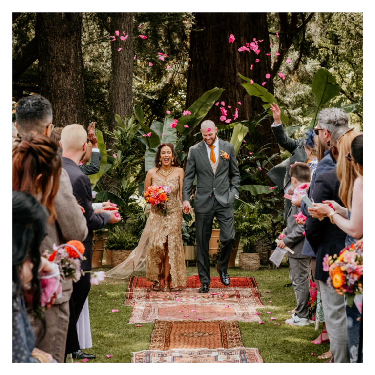 Newlywed couple walking back down the aisle as guests throw pink petal confetti in an outdoor woodland wedding ceremony