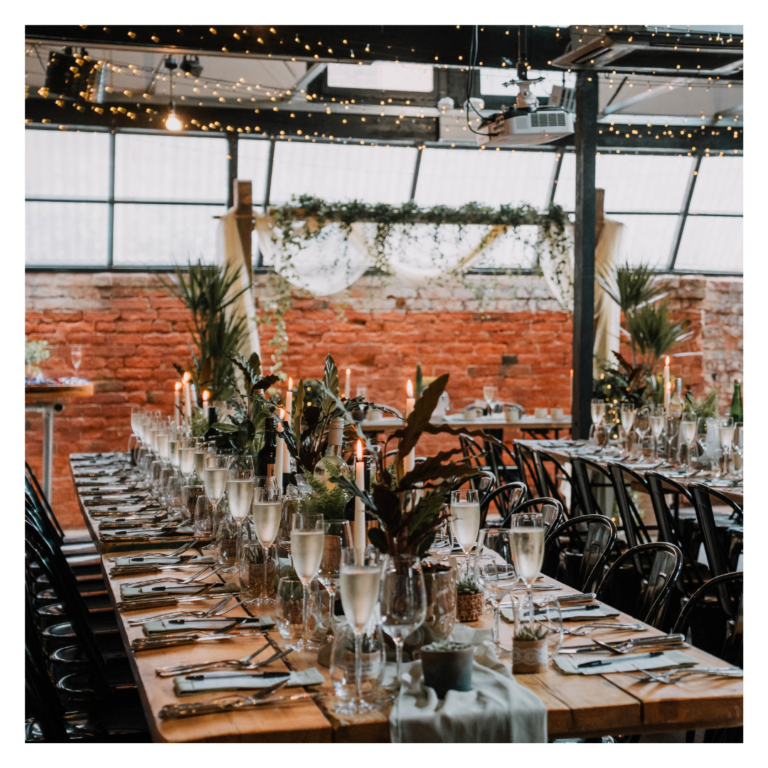 Long wooden wedding tables styled with greenery, candles and glassware in an industrial venue
