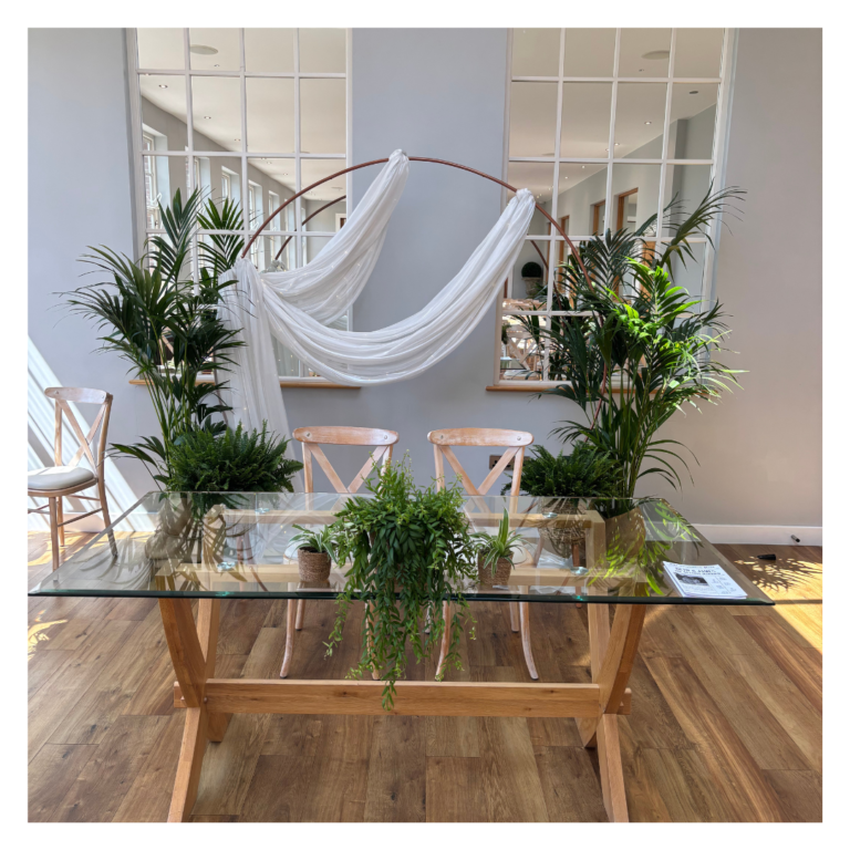 Wedding ceremony setup with rose gold arch, white draping, greenery and glass signing table.