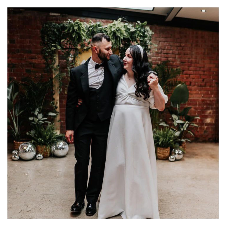 Bride and groom walking together beneath a greenery arch with disco balls and brick backdrop