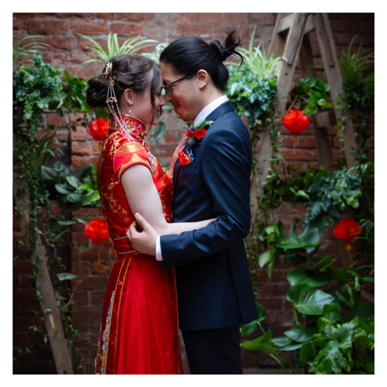 Couple embracing in front of a greenery wedding backdrop with red lantern accents