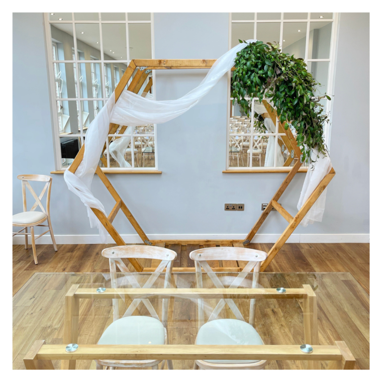 Hexagon wedding arch with greenery and white draping positioned behind ceremony table