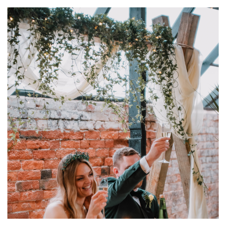 Bride and groom raising a toast beneath a greenery and draped wedding head table backdrop