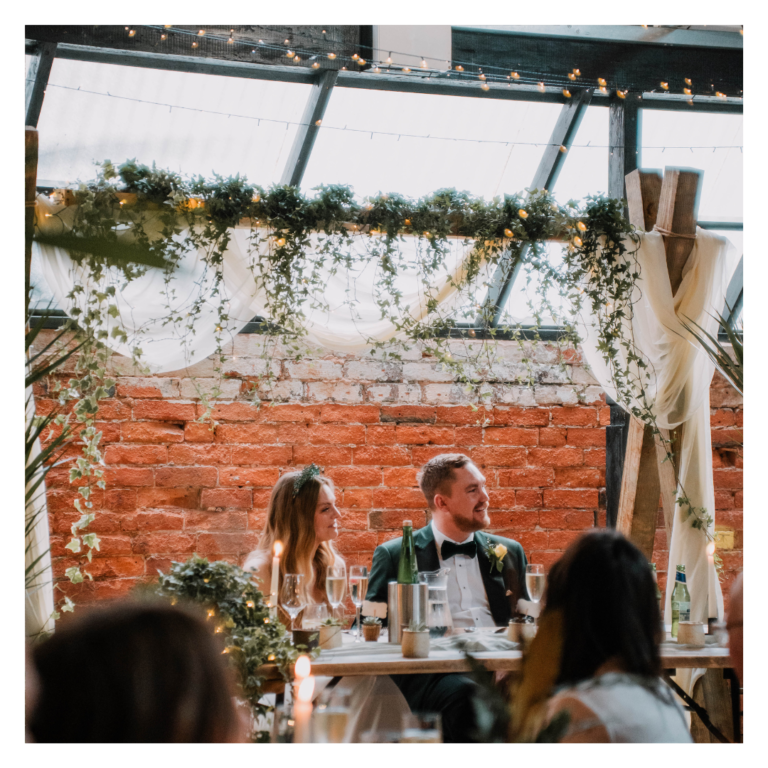 Wedding top table beneath a greenery ceremony backdrop with fairy lights and white draping
