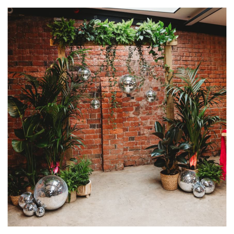 Lush greenery wedding arch with hanging disco balls against an exposed brick wall backdrop