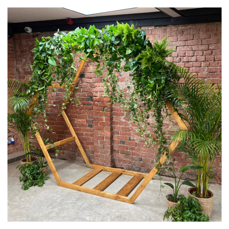 Angled view of a hexagon greenery wedding arch framed by potted palms and trailing foliage