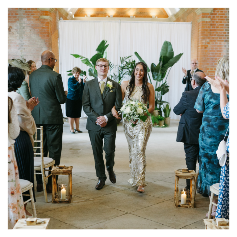 Bride and groom walking down the aisle with greenery backdrop and candle lanterns.
