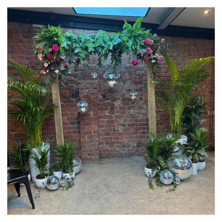 Floral greenery wedding arch with pink roses, disco balls and potted plants against a brick wall