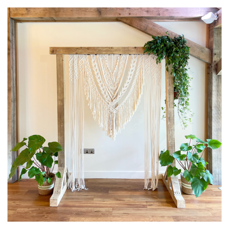 Boho wedding ceremony backdrop with macramé hanging, wooden frame and trailing greenery.