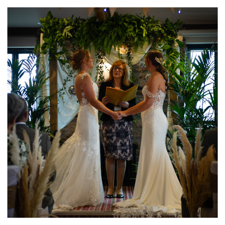 Same-sex wedding ceremony beneath a greenery arch with draped fabric and hanging disco balls