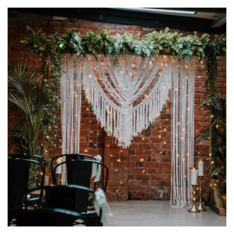 Boho macramé wedding backdrop with fairy lights and greenery against a brick wall