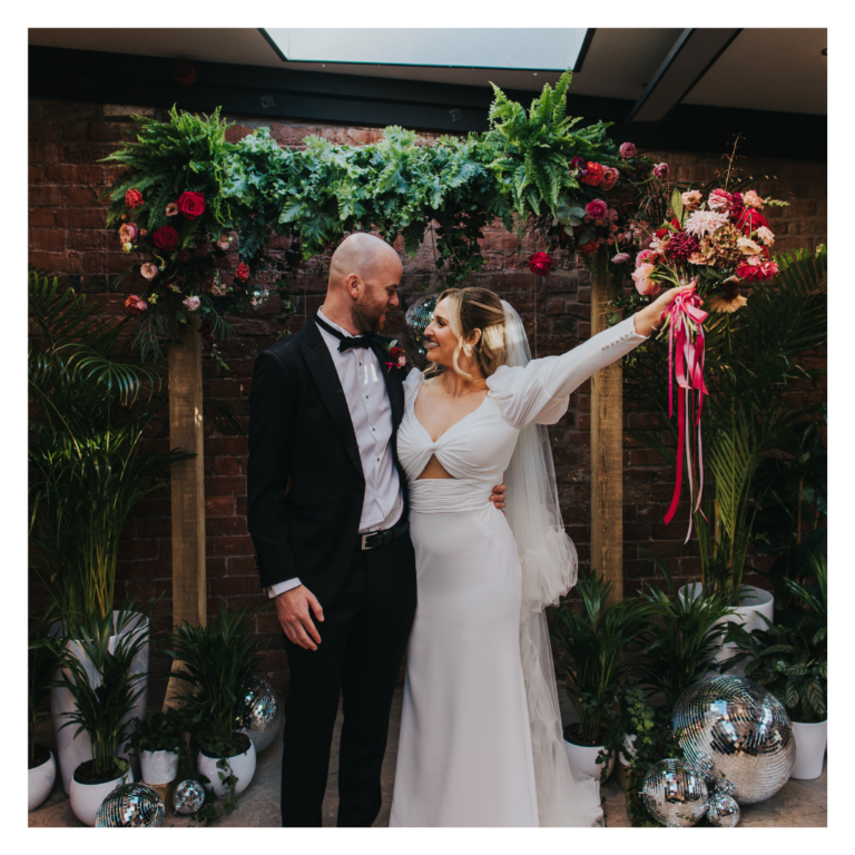Bride and groom standing beneath a lush botanical ceremony backdrop with greenery, florals and disco ball accents at an indoor wedding venue.