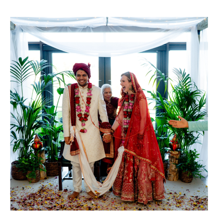 Bride and groom laughing during a Hindu wedding ceremony beneath a botanical mandap with white draping and greenery