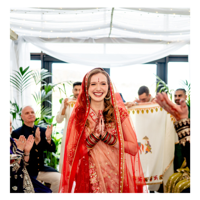Bride entering a Hindu wedding ceremony beneath a botanical mandap with white draping and greenery