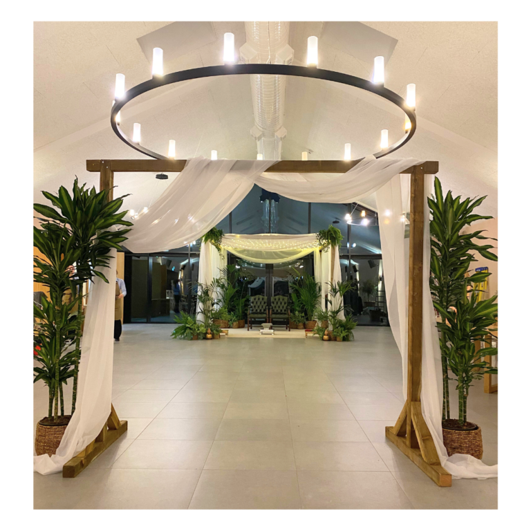 Botanical mandap entrance with wooden frame, white draping and greenery leading to a Hindu wedding ceremony space