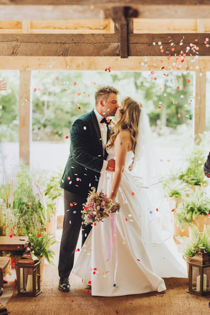 Bride and groom kissing during their wedding ceremony as colourful confetti falls, surrounded by greenery and sustainable décor in a rustic UK wedding venue.
