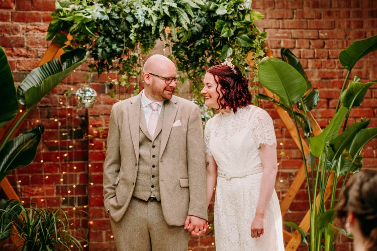 Married couple infront of a hexagon backdrop with plants and disco balls. The Bride is wearing a gorgeous jumpsuit